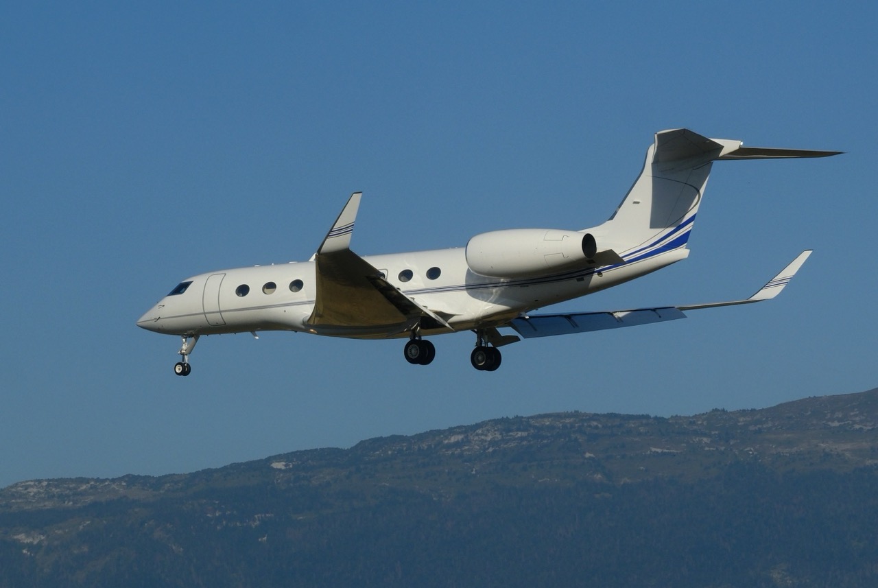 Gulfstream G650ER private jet landing at Geneva Airport with landing gear down and mountain backdrop.