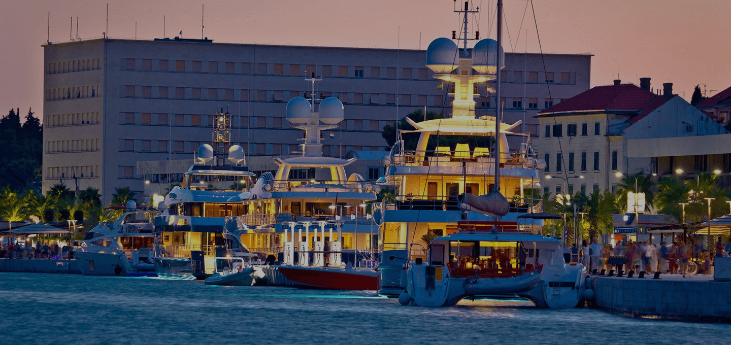 Super yachts docked at a marina for a yacht show at sunset with visitors looking at the boats.